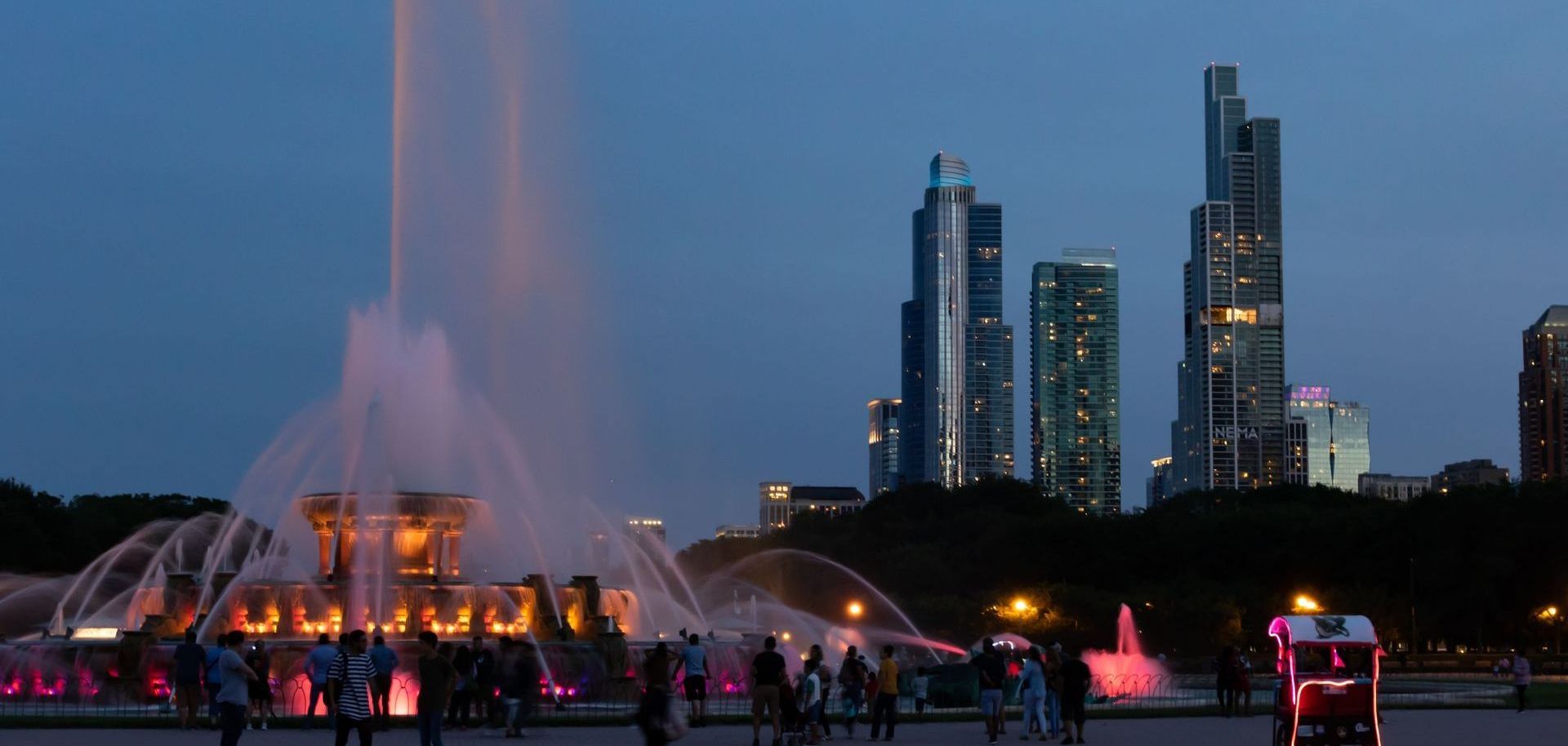 A fountain in a park with a city skyline in the background at night.