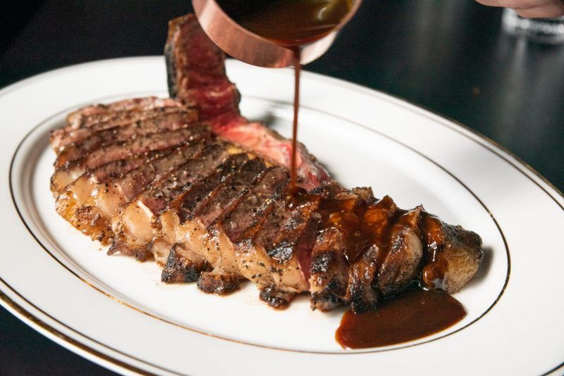 A person is pouring sauce on a steak on a white plate.