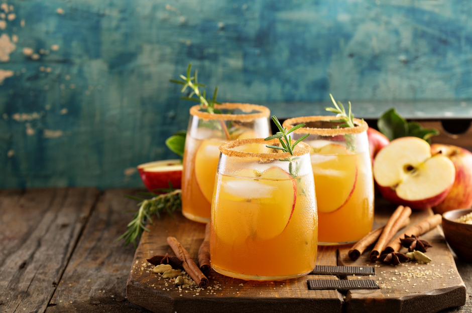 Three glasses of apple cider with ice and cinnamon sticks on a wooden cutting board.