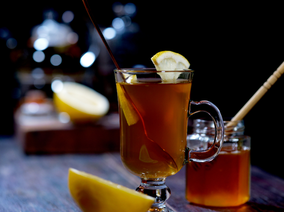 A glass of tea with a slice of lemon and honey next to a jar of honey.