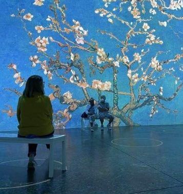 A woman sits on a bench in front of a painting of a tree with flowers