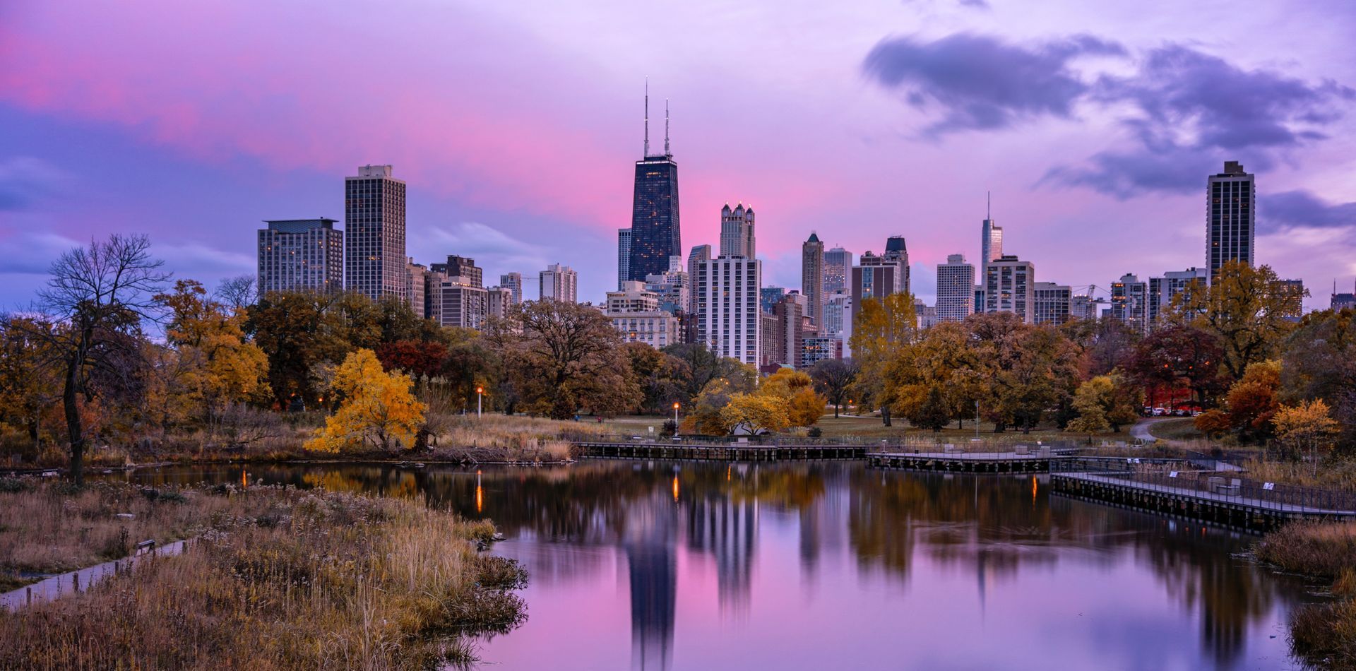 A city skyline is reflected in a lake at sunset.
