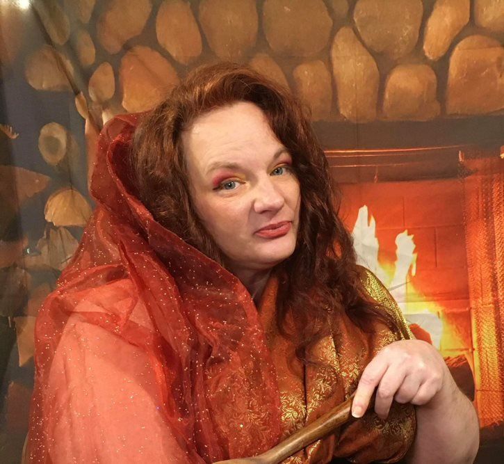 A woman is holding a wand in front of a fireplace,  representing Hestia.