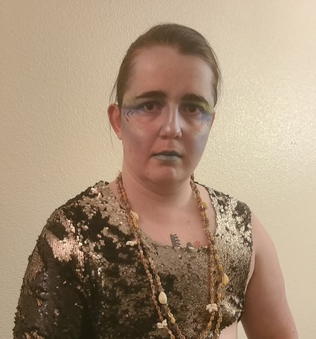 Someone wearing a sequined top and necklace is standing in front of a white wall, representing Poseidon. 