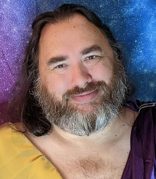 A man with a beard and long hair is smiling for the camera, representing Zeus.