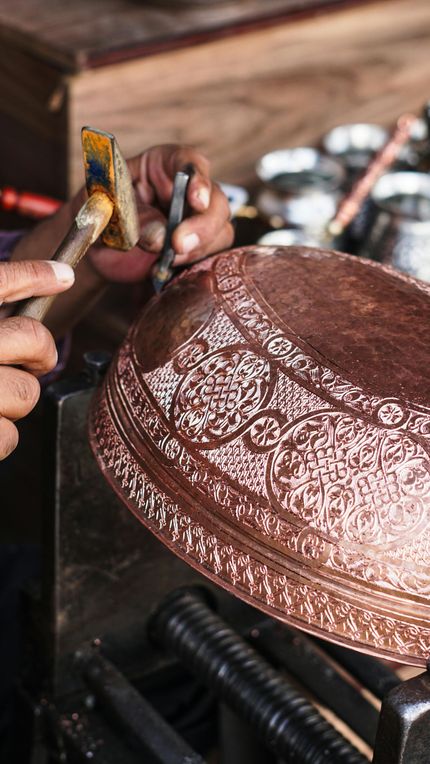Hands hammering a copper pot on a forge, shaping the hammered surface.
