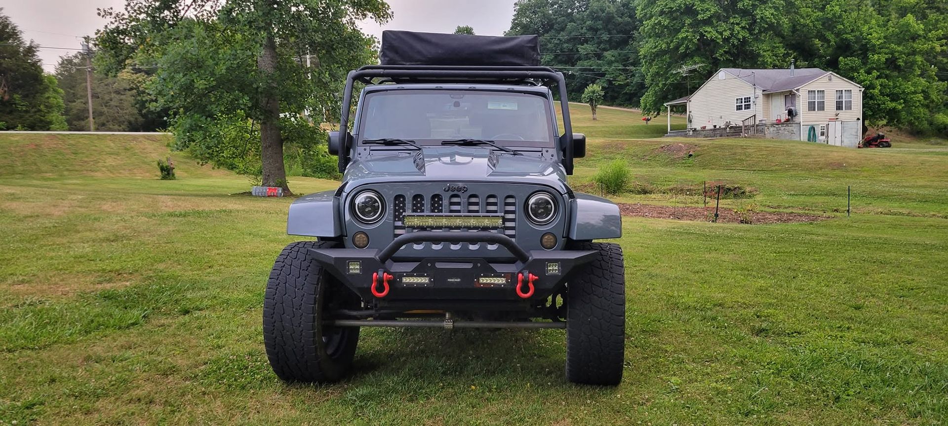Gray off-road Jeep on grass with roof cargo rack, parked in a rural yard near trees and a white house.
