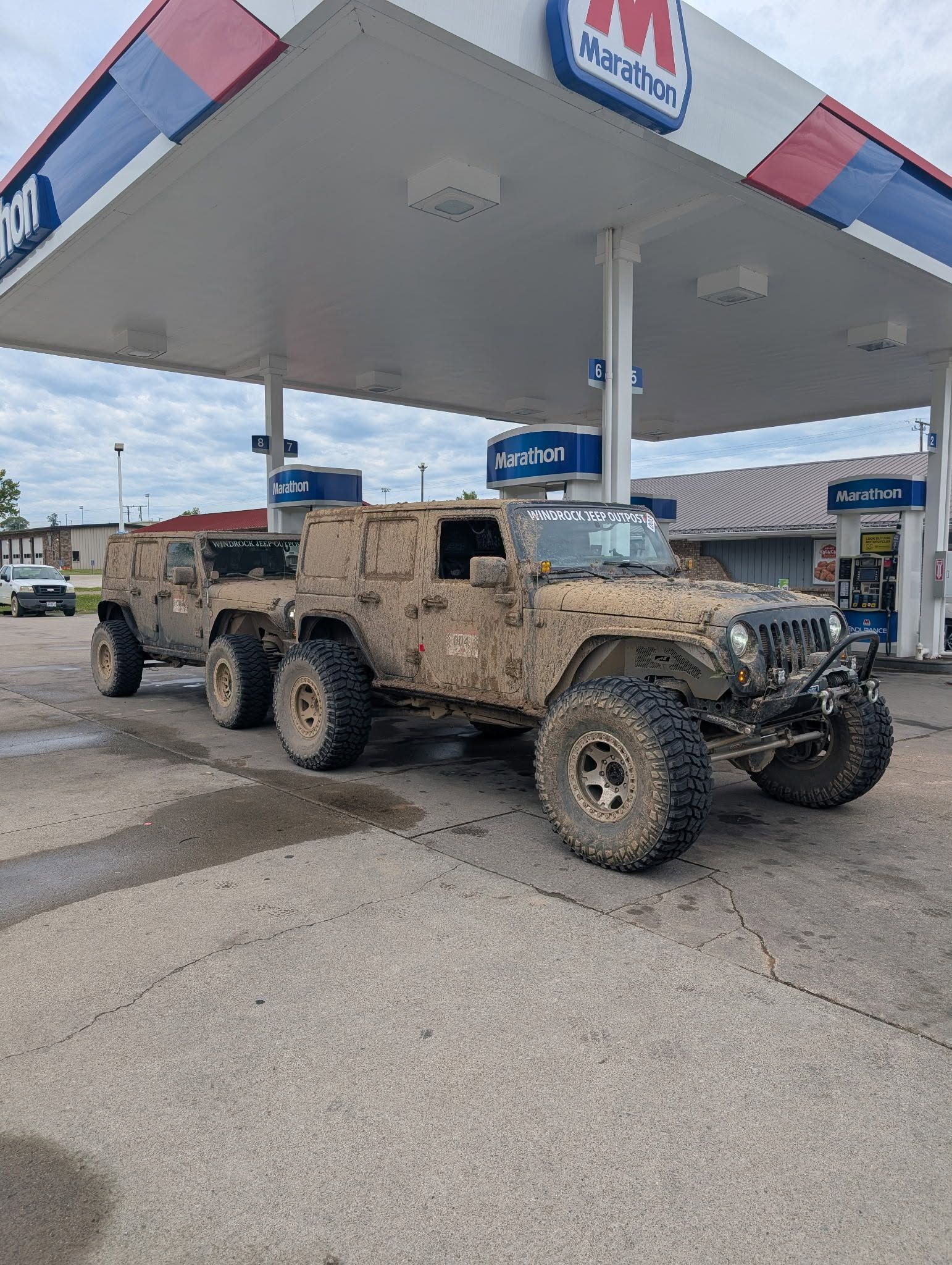Camouflaged military vehicle parked under a gas station canopy on a wet pavement