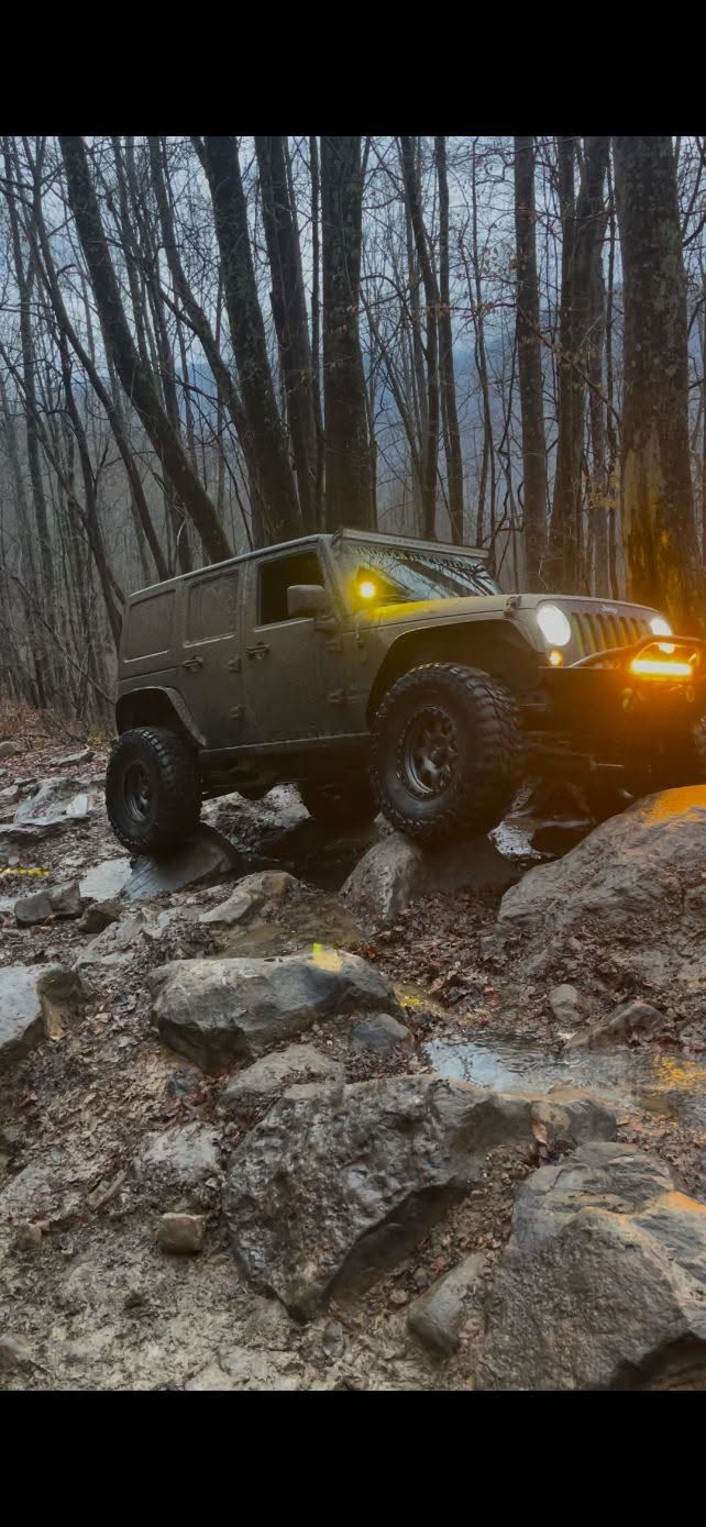 Lifted off-road Jeep crawling over rocks in a snowy forest at dusk, headlights glowing.