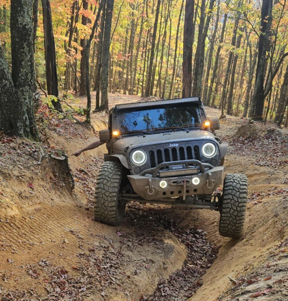 Black Jeep Wrangler crawling through a muddy forest trail with headlights on
