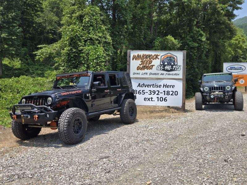 Two black off-road Jeeps parked by an advertising sign on a gravel lot surrounded by trees