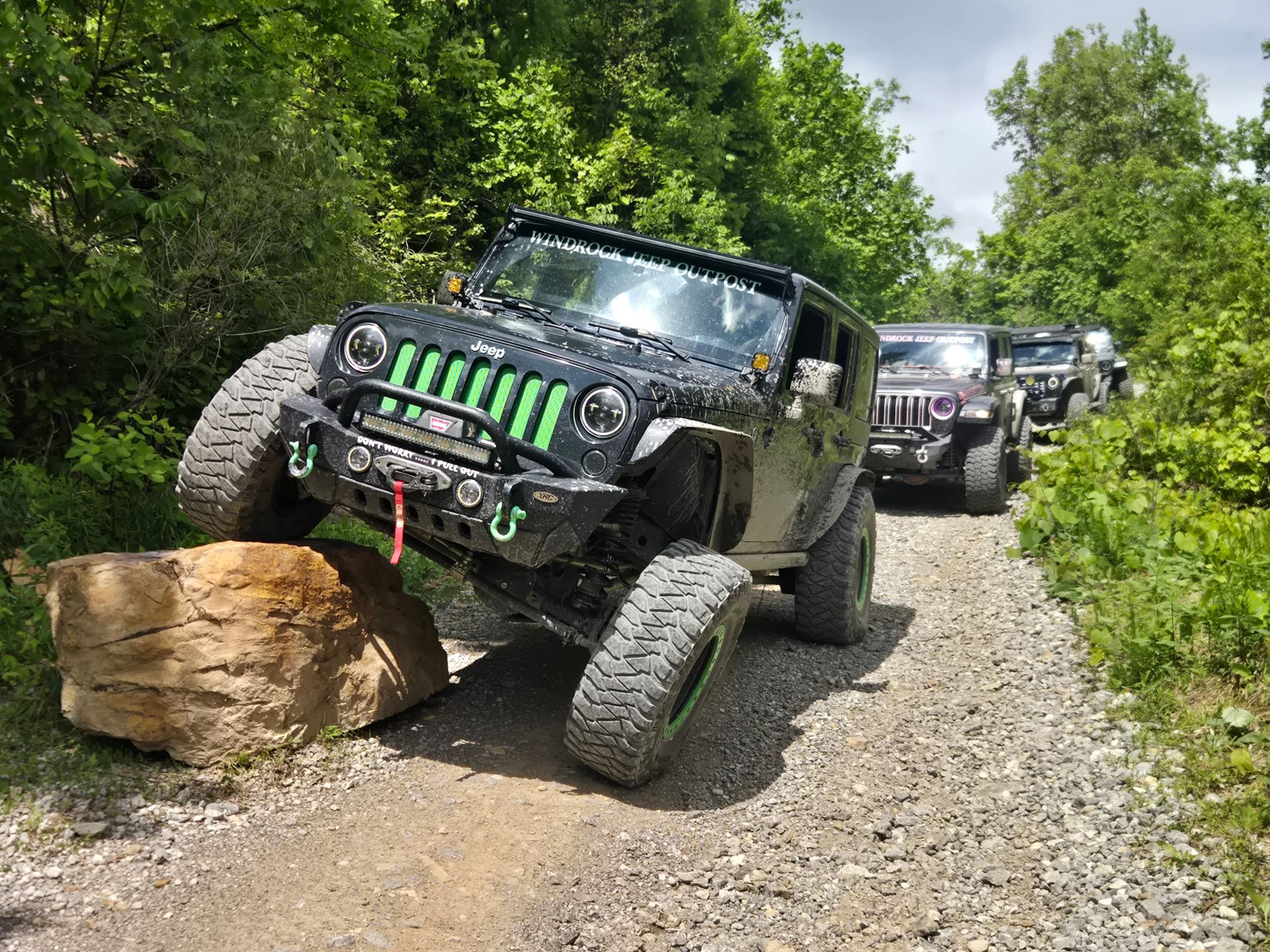 Black Jeep off-roading over a boulder on a forest trail, with other vehicles behind.