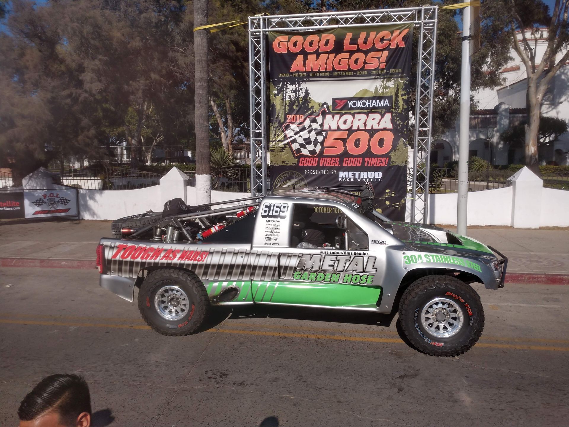 Green and white off-road race truck at the NORRA 500 starting line under a “Good luck amigos!” banner