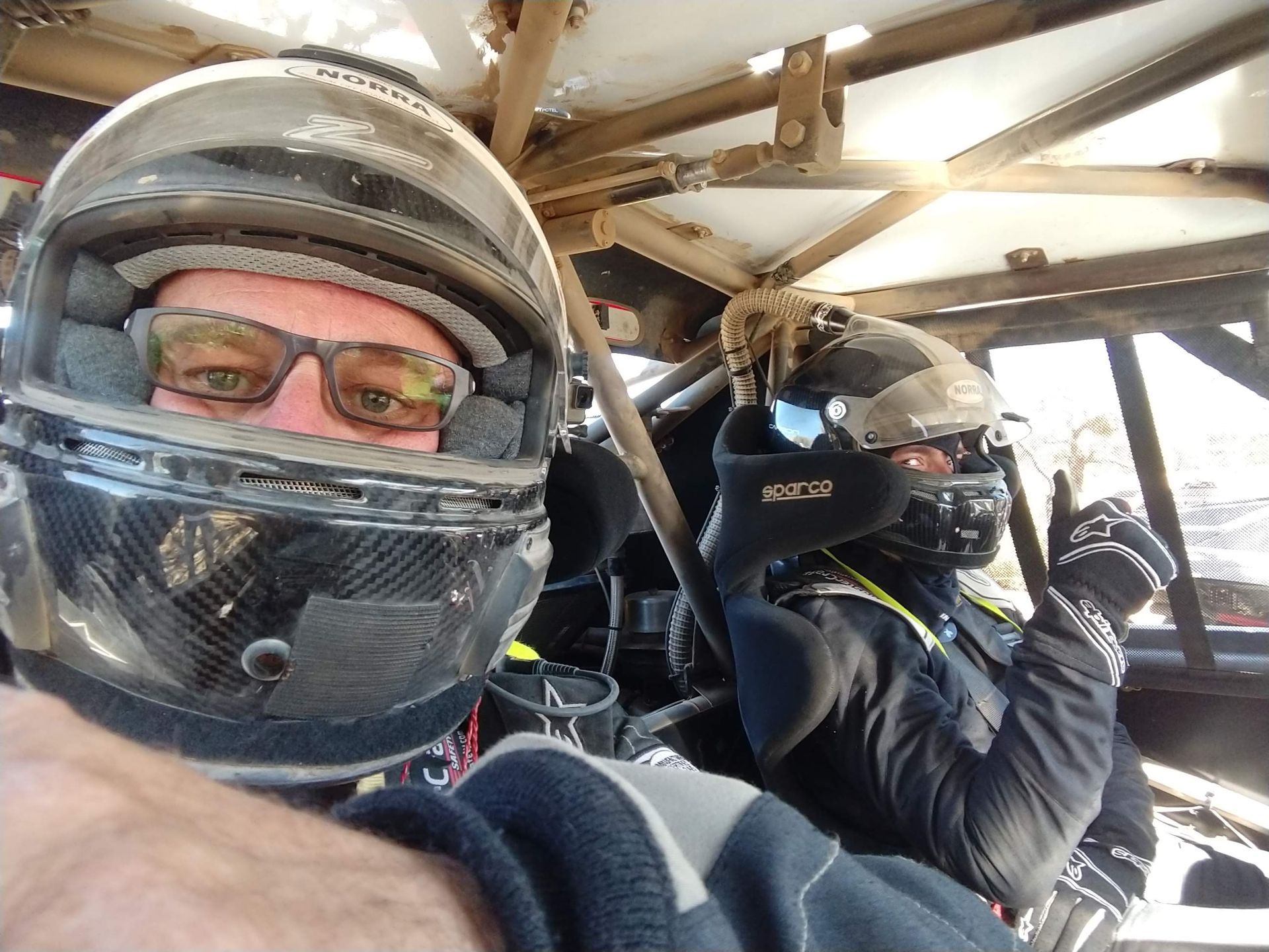 Two helmeted off-road racers in a vehicle, one taking a selfie inside the roll cage