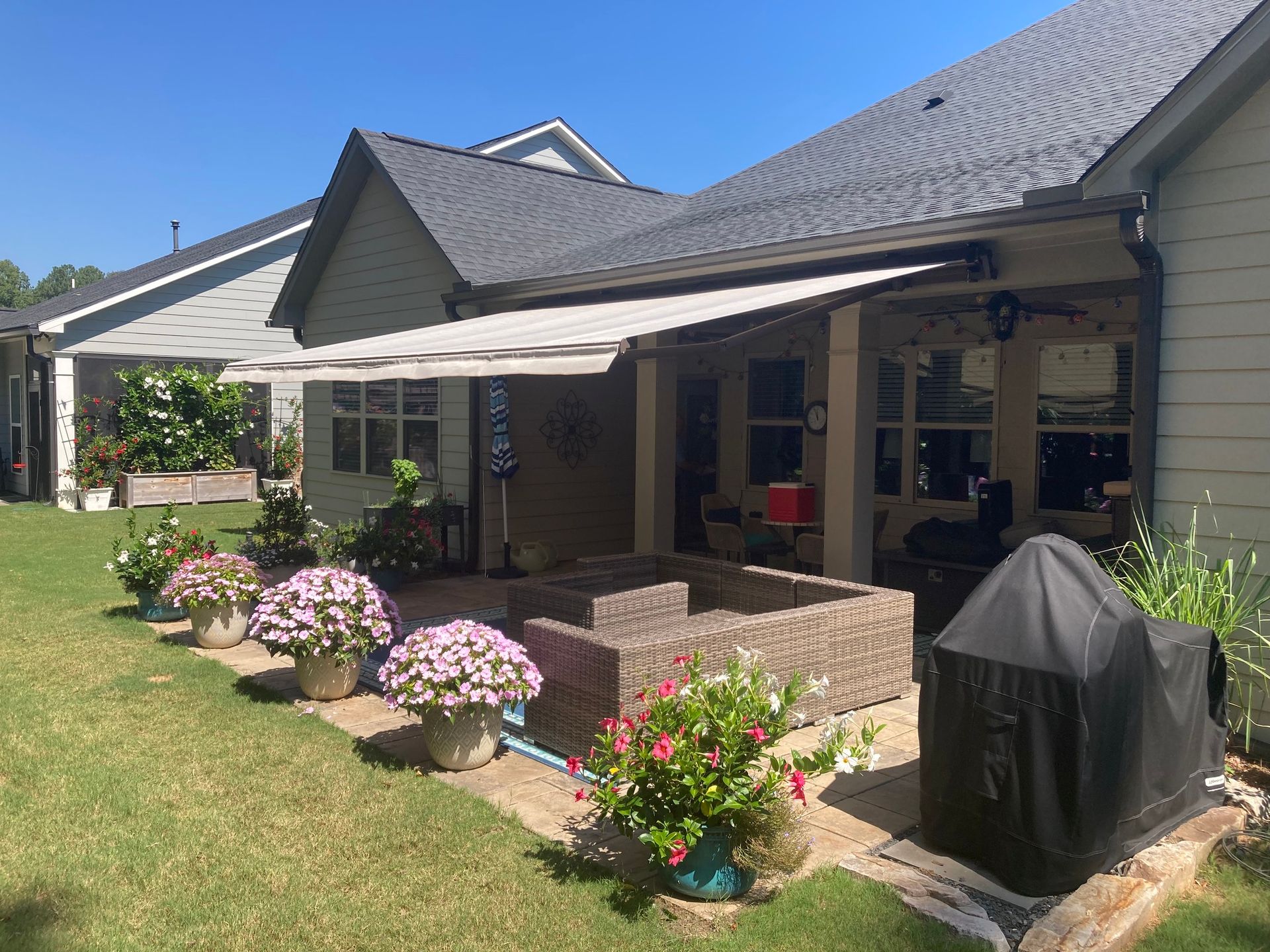 Patio with tan awning, flowers in pots, wicker furniture, and covered grill.
