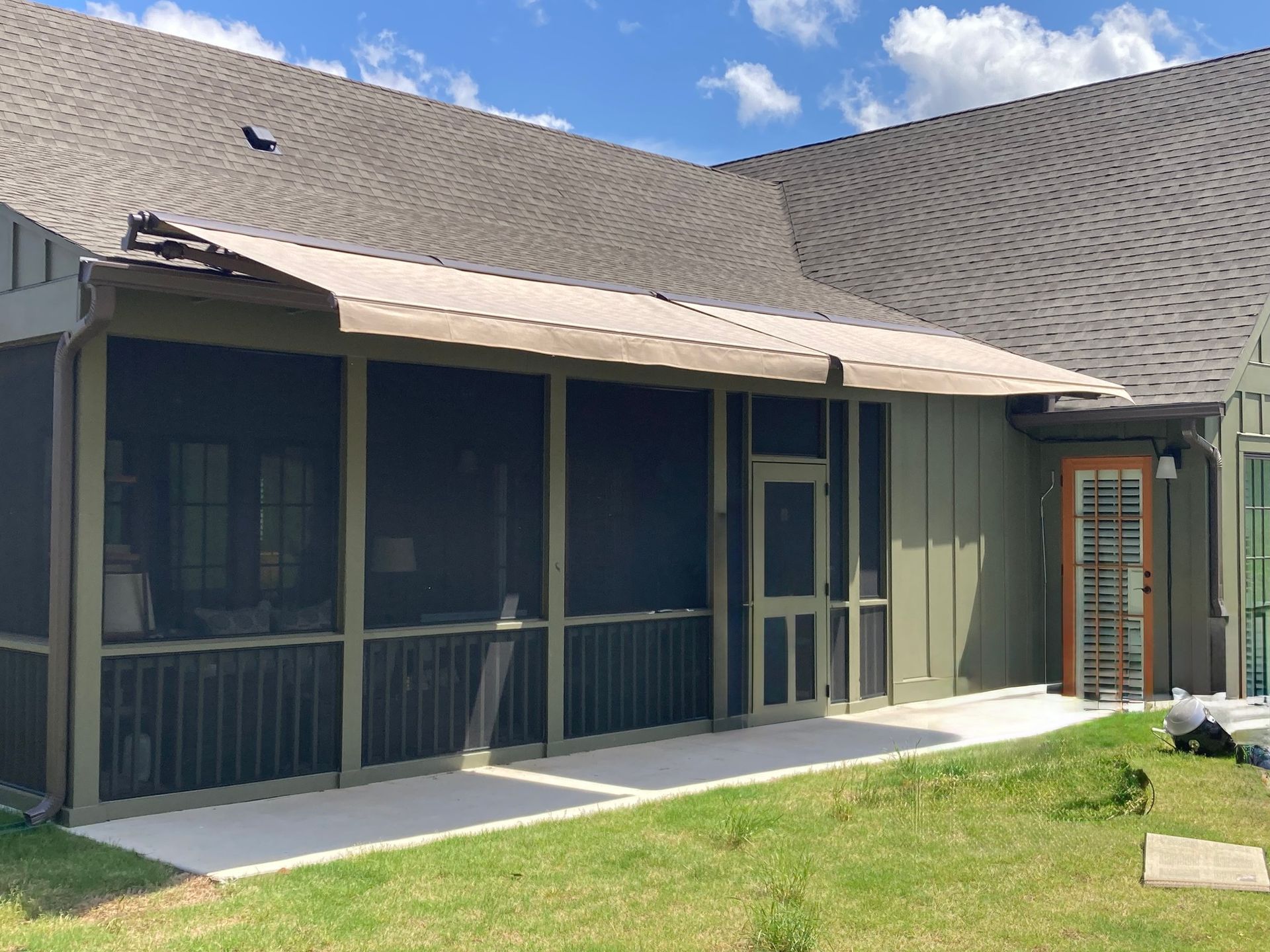 Screened porch with retractable awning on a sunny day. Beige awning, green siding, and a concrete patio.