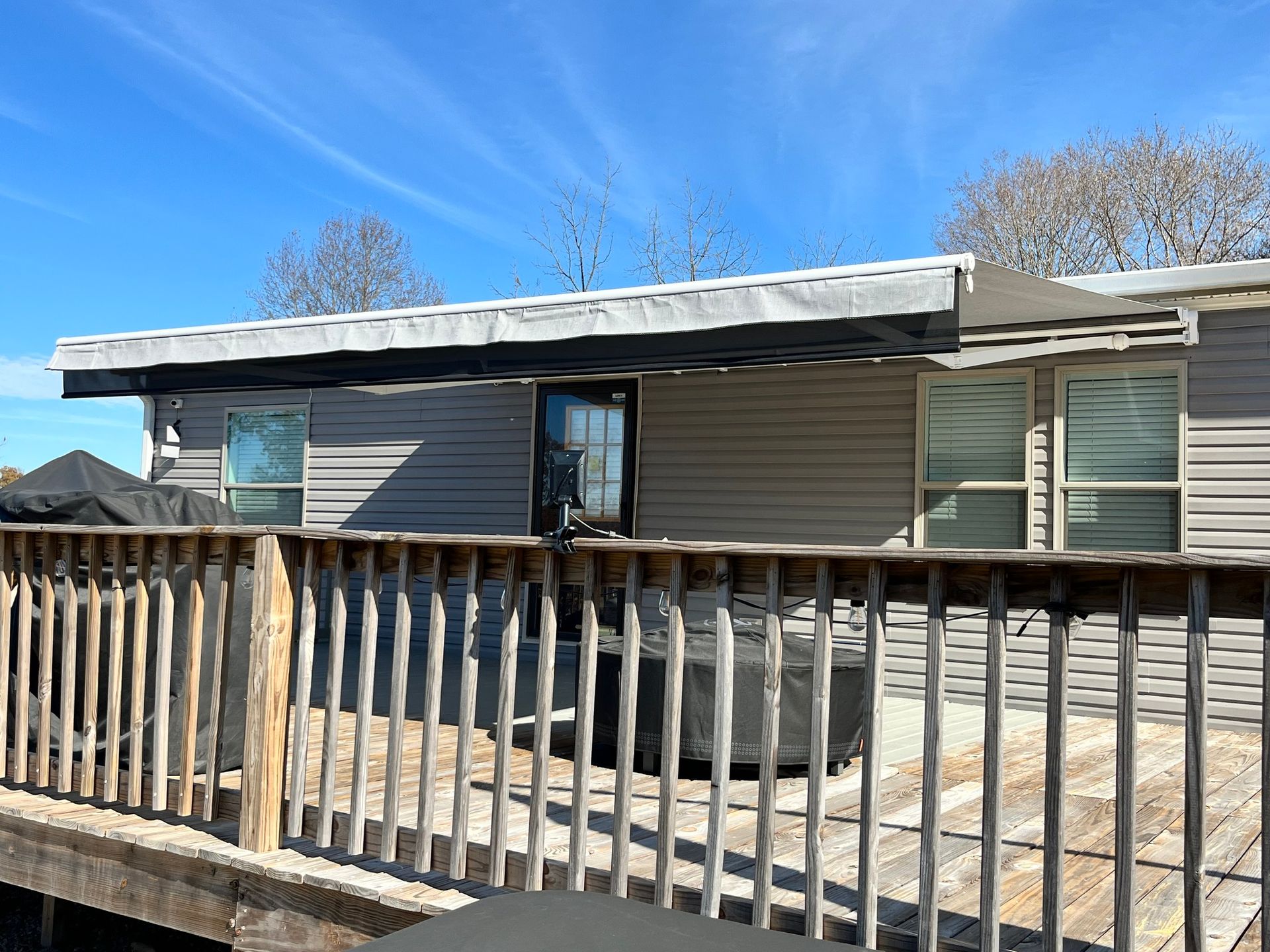 Backyard deck with gray siding house, awning, windows, and wooden railing under a blue sky.