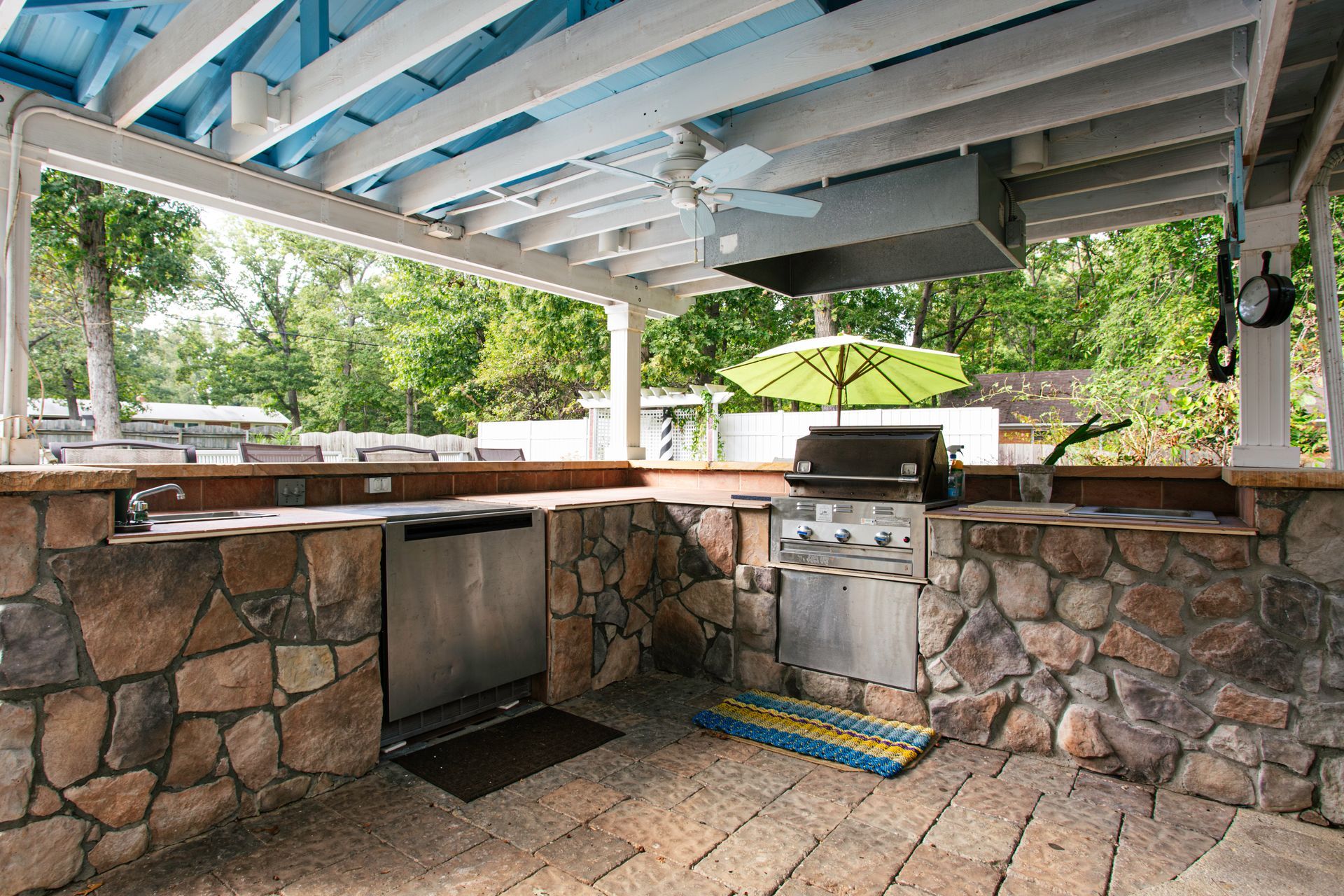 Outdoor kitchen with stone facade, grill, sink, and shaded by a blue-painted pergola.