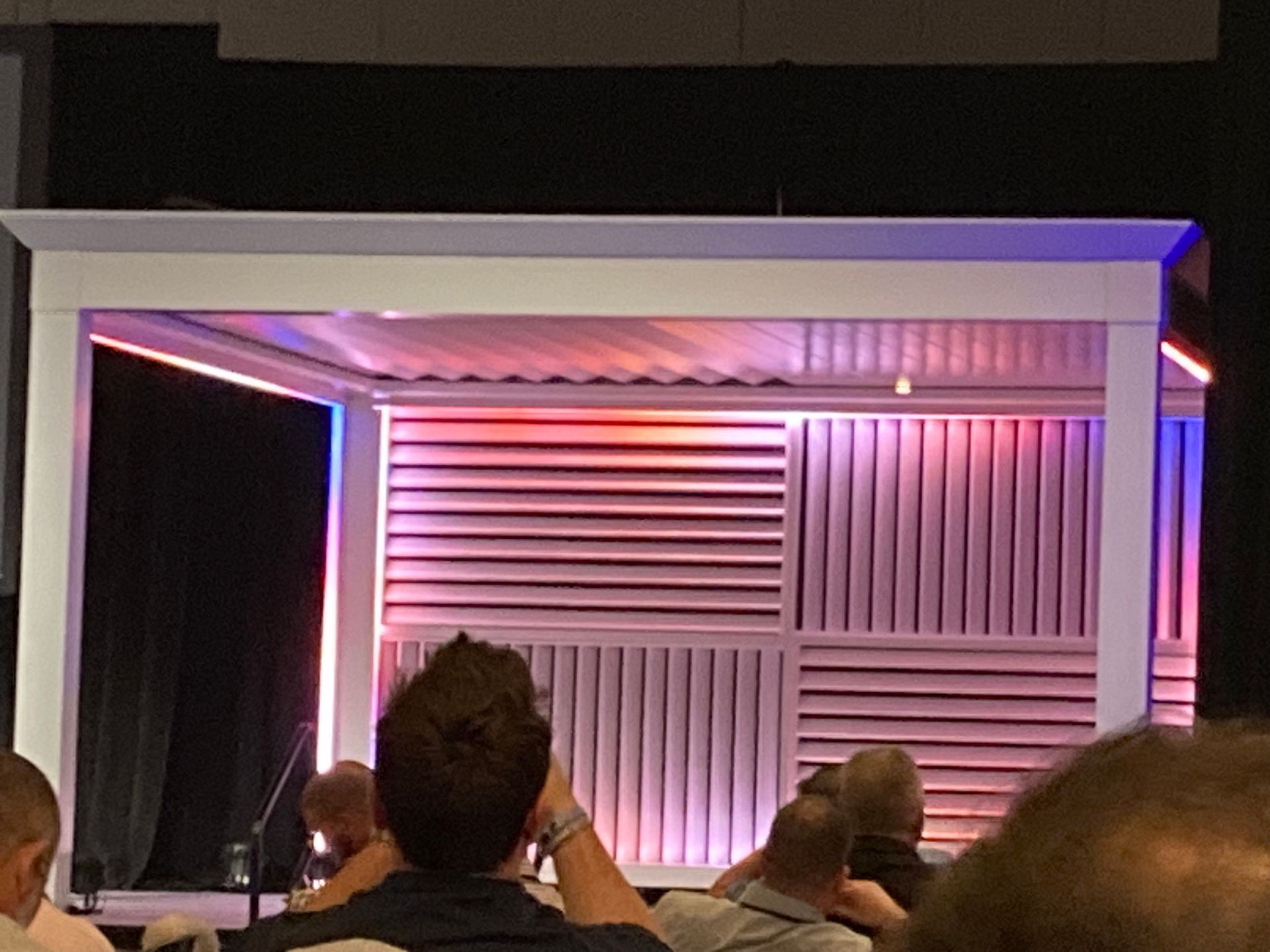 Stage with white frame, wooden slat backdrop, and colorful lighting. People seated in foreground.