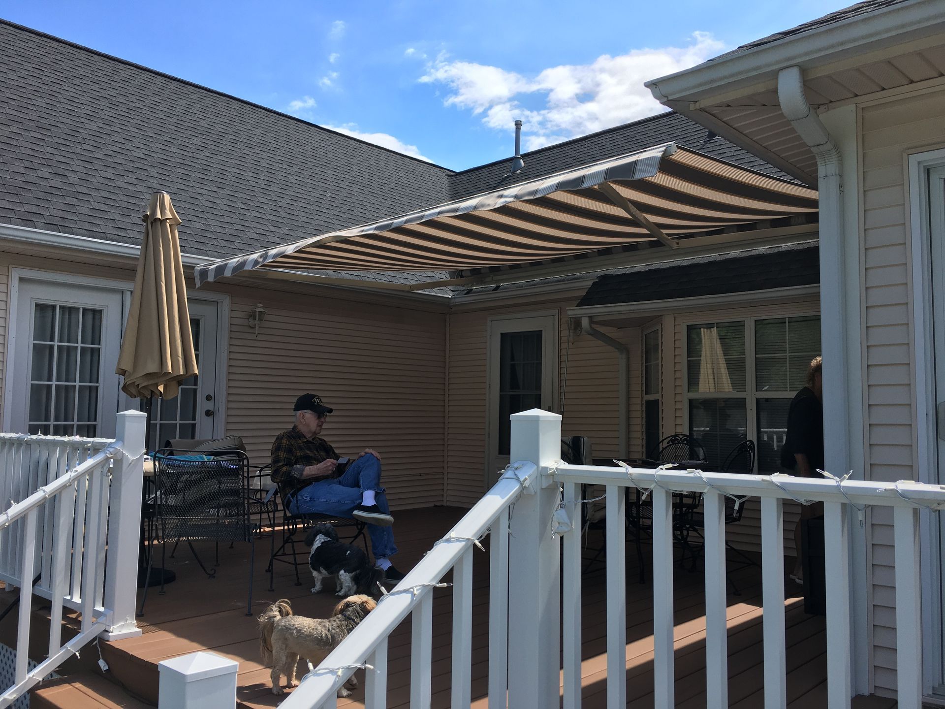A person sitting on a patio under a striped awning, a dog, and a deck with a white railing.