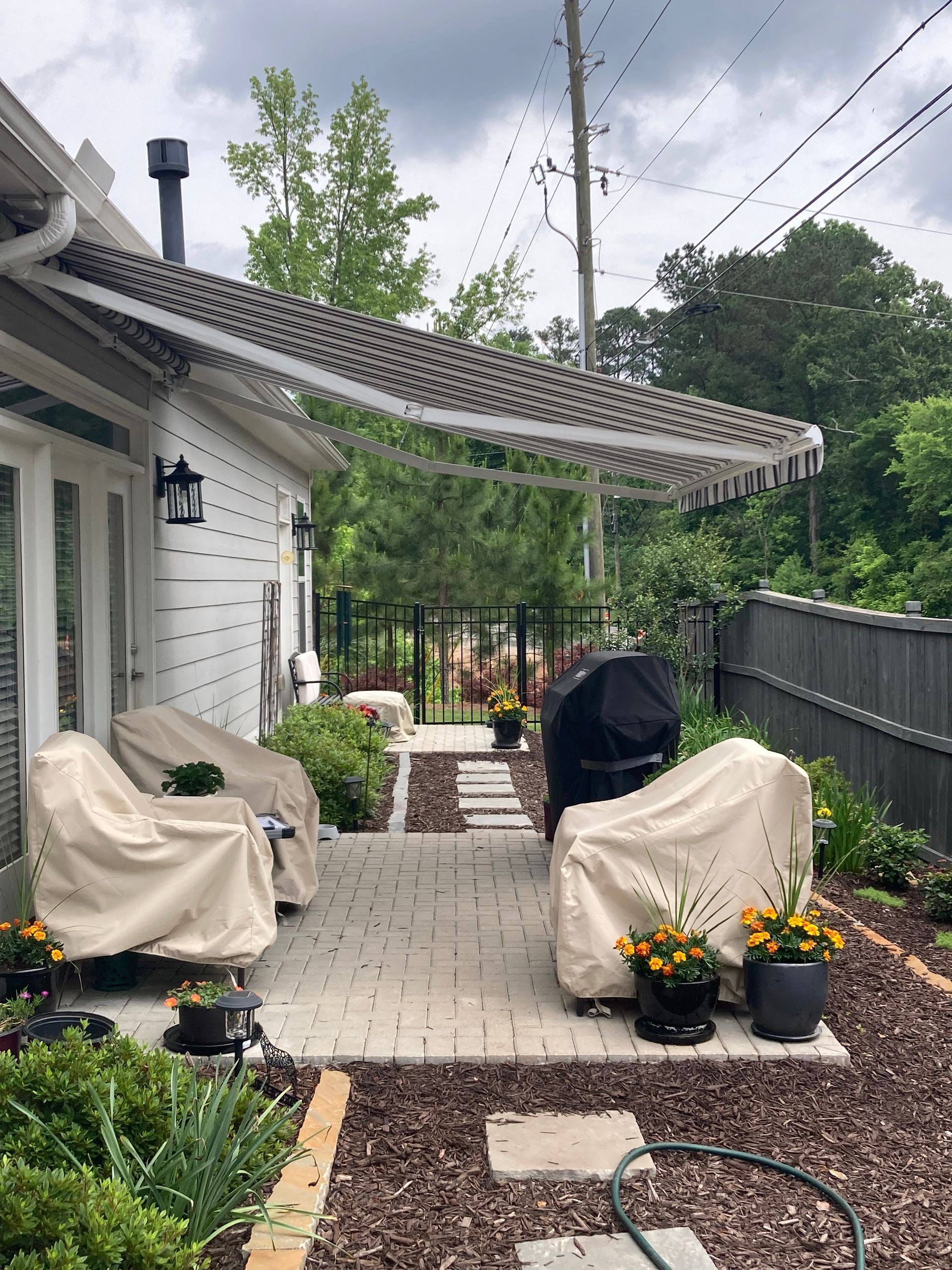 Outdoor patio with covered chairs, grill, and walkway surrounded by plants.