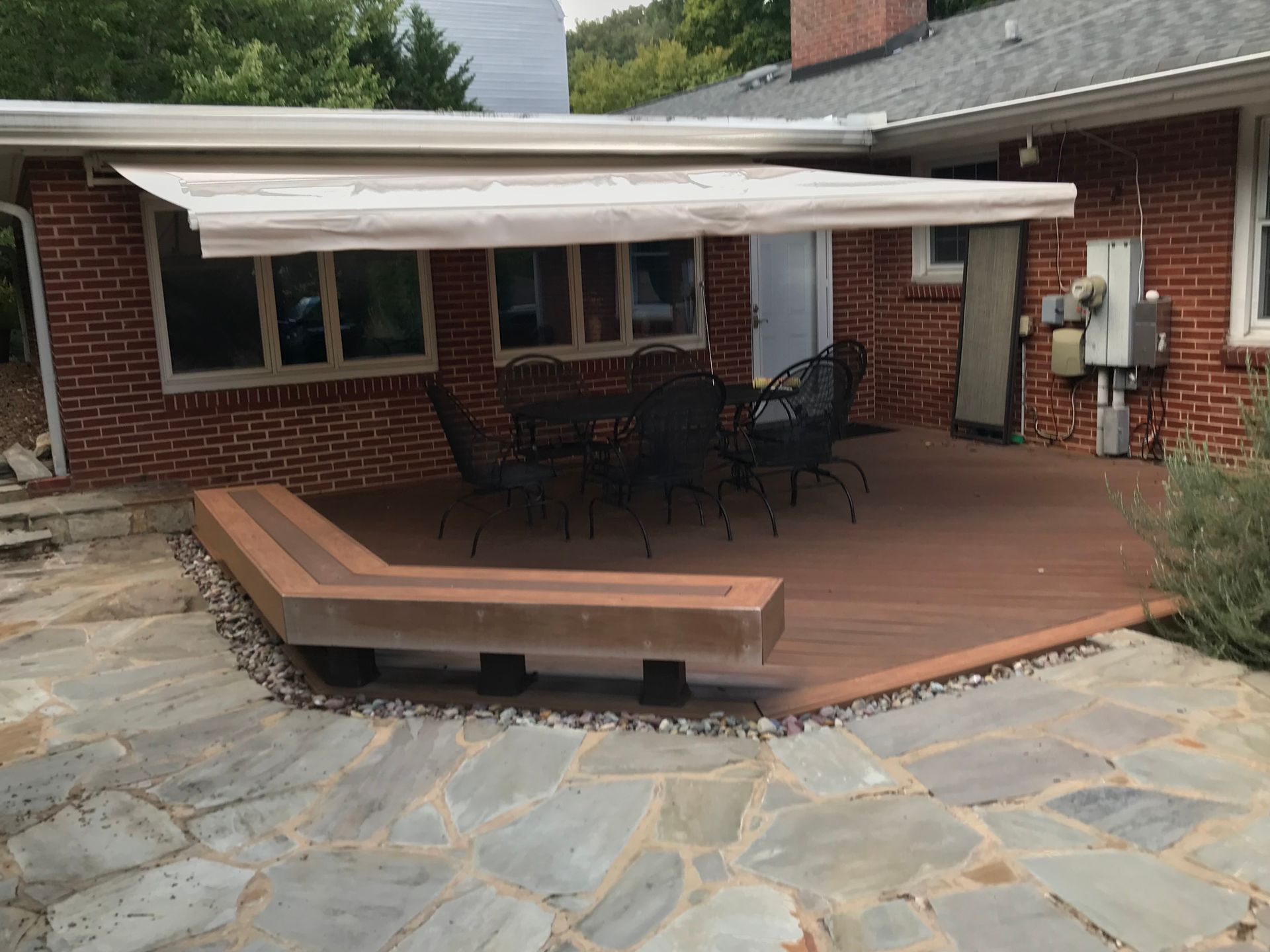 Deck with awning, seating, and a table in front of a brick house. Stone patio surrounds the deck.