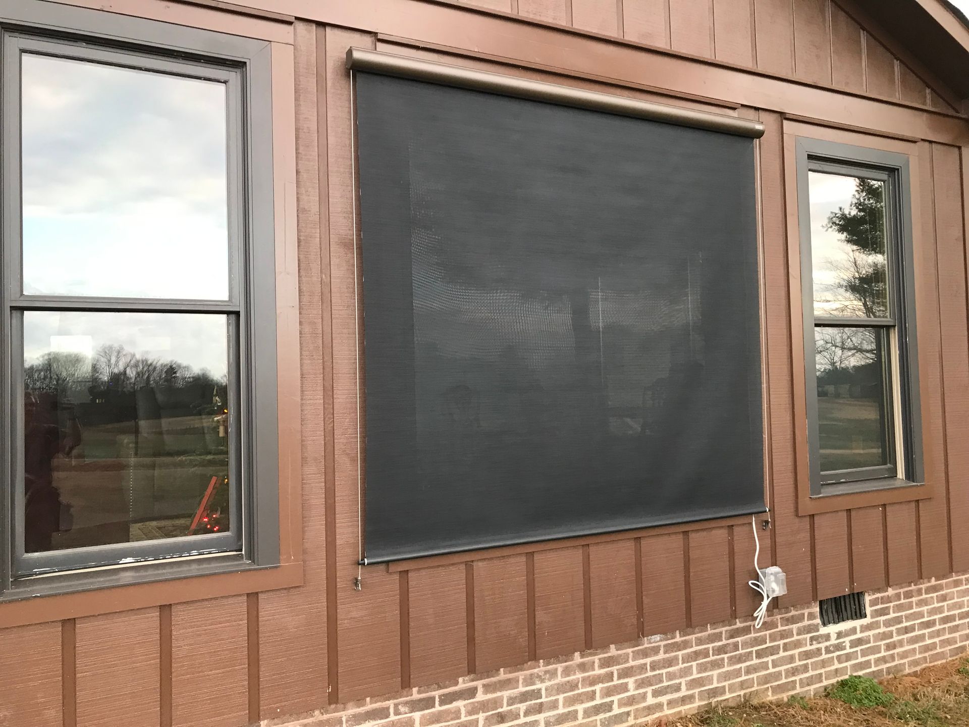 Brown house exterior with dark screen shade over a window, flanked by two other windows with dark frames.