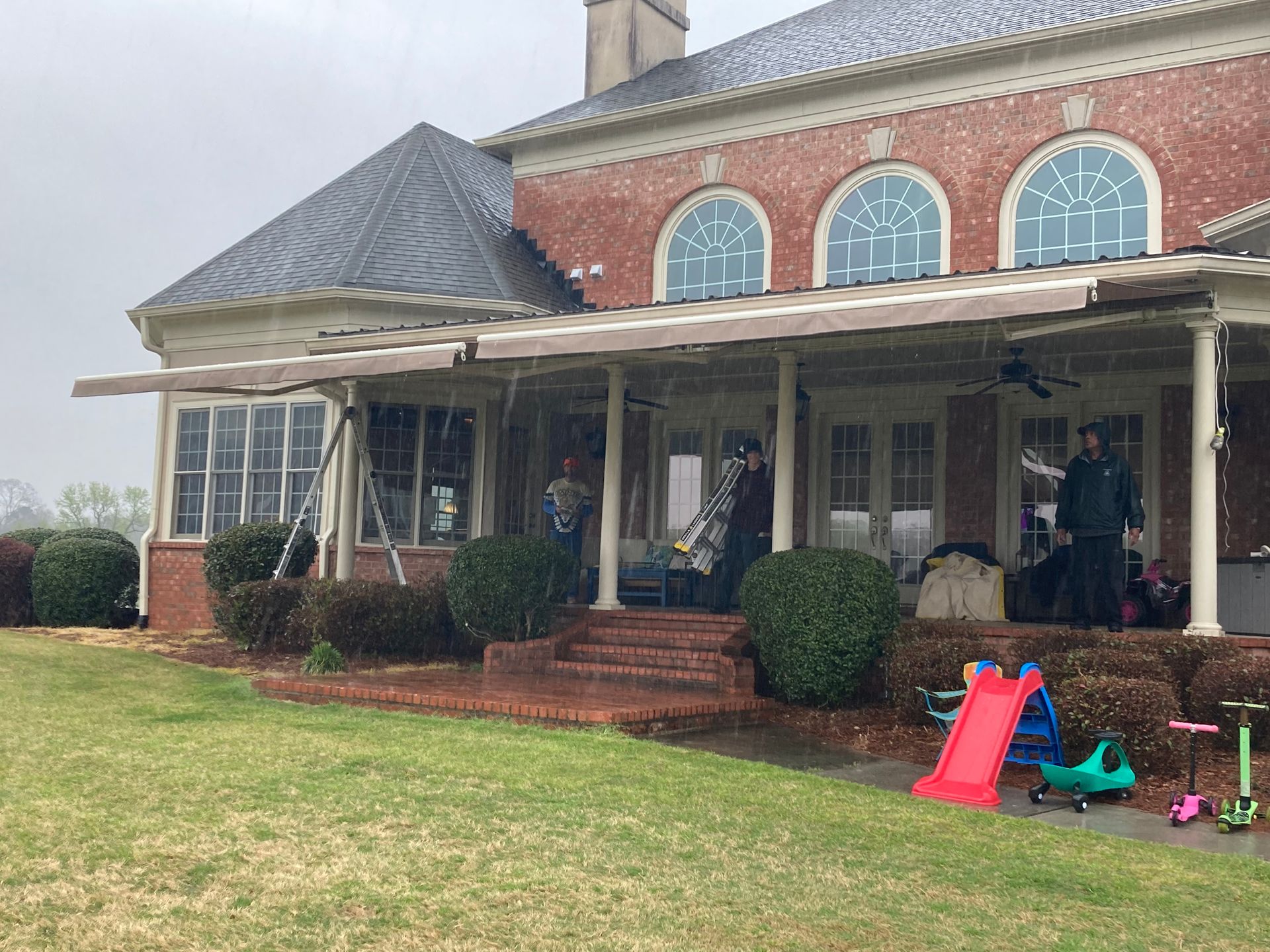 Rainy day view of a brick house with a covered porch. People stand on the porch and yard, along with kids' toys.