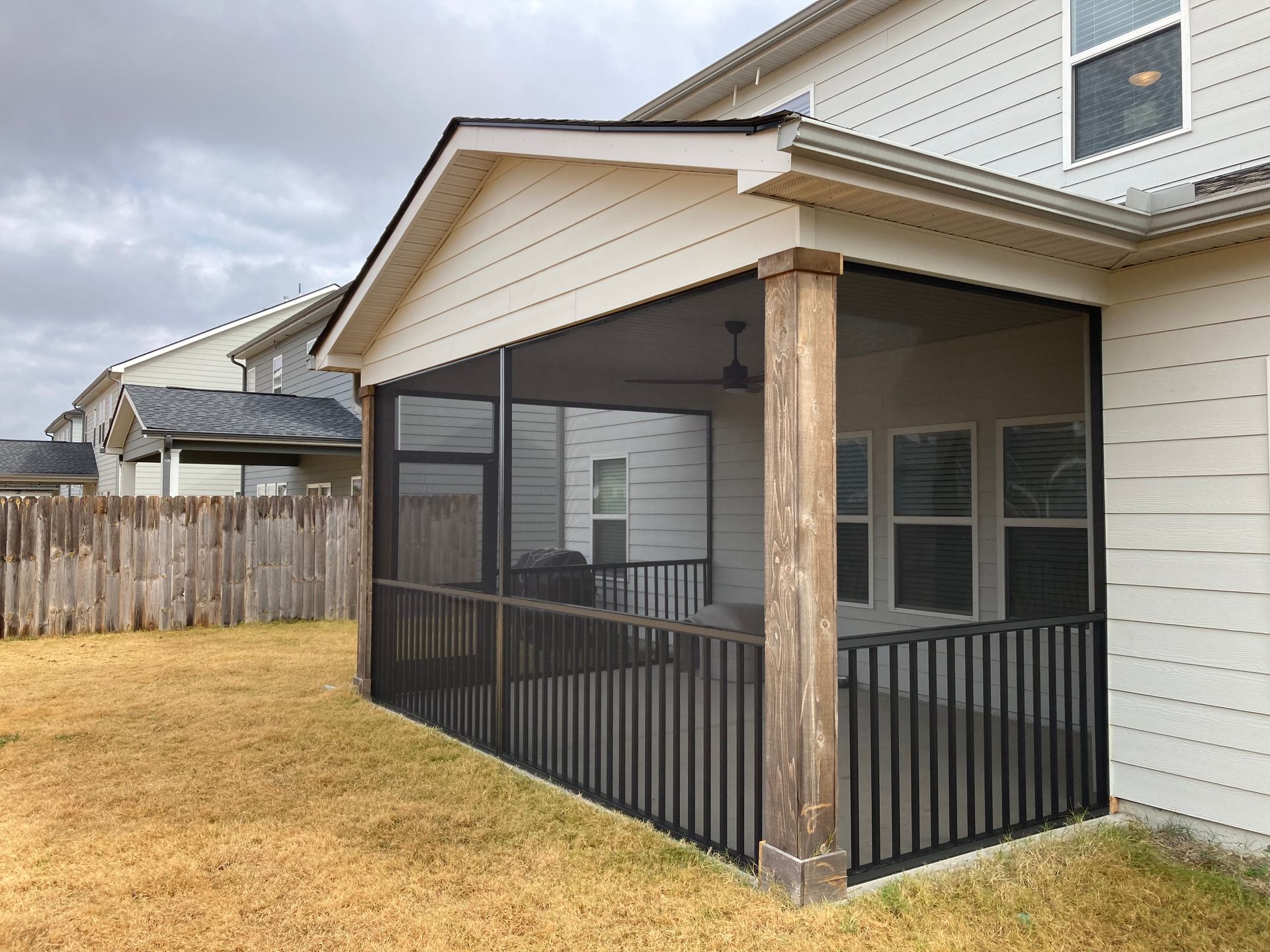Screened-in porch on a house with brown wooden posts and black railings. Brown grass and cloudy sky.