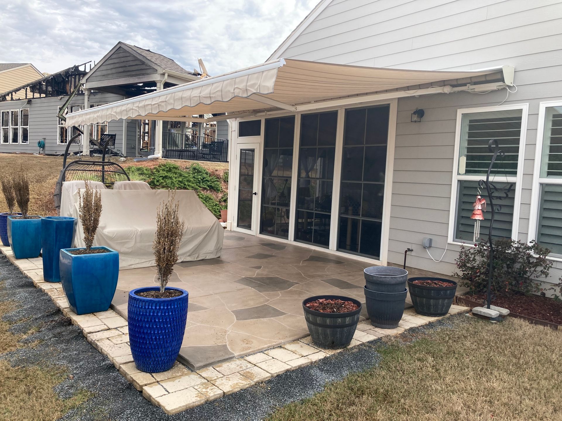 Patio with awning, screened porch, potted plants, and partially visible damaged house in the background.