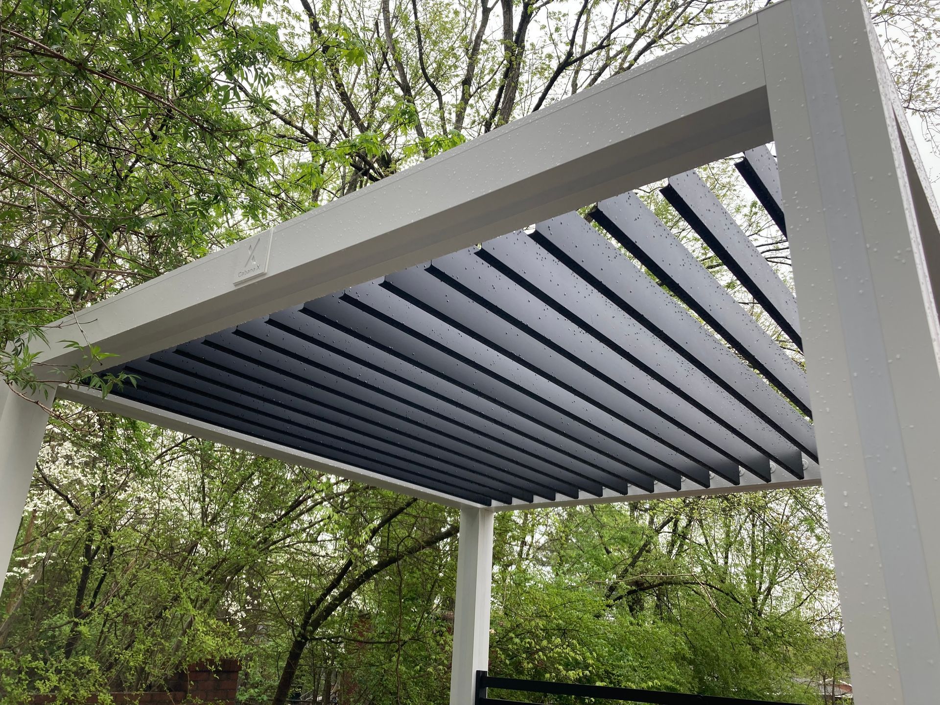 White pergola with dark gray adjustable louvers, set against a background of green trees.