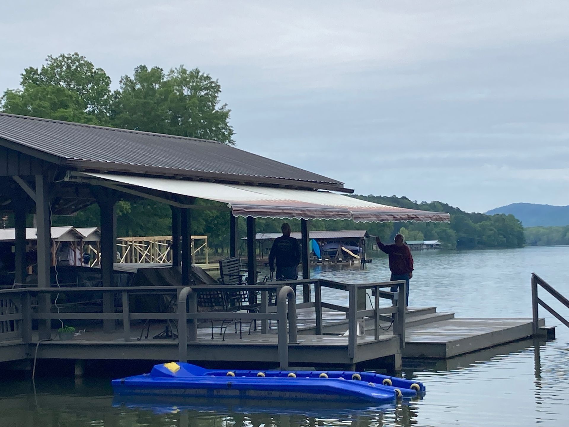 Two people stand on a wooden dock by a lake, beneath a covered structure. One waves. A blue watercraft floats in the water.