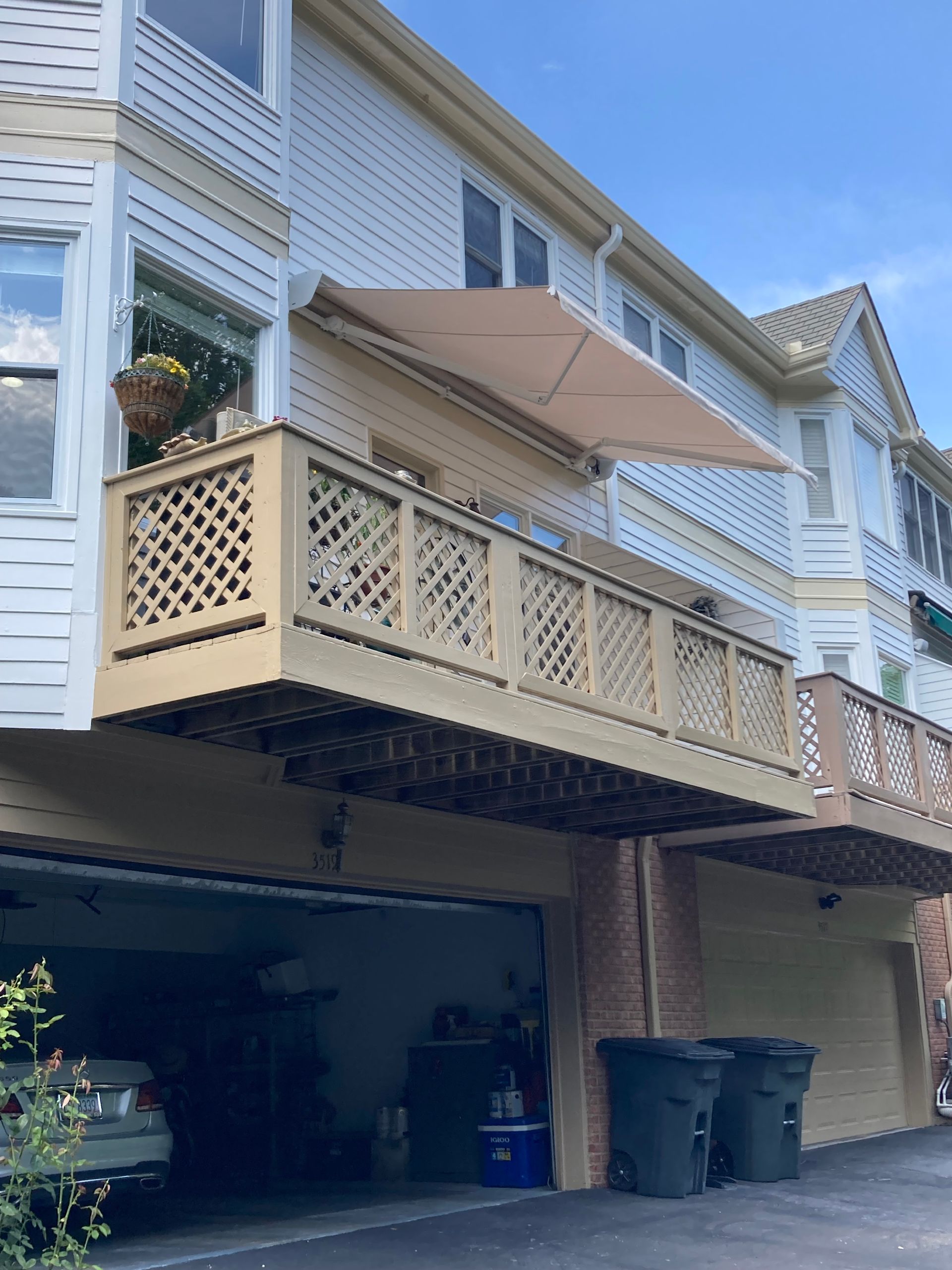 Beige awning extends over a wooden deck, part of a townhouse complex. Garage below, blue sky.