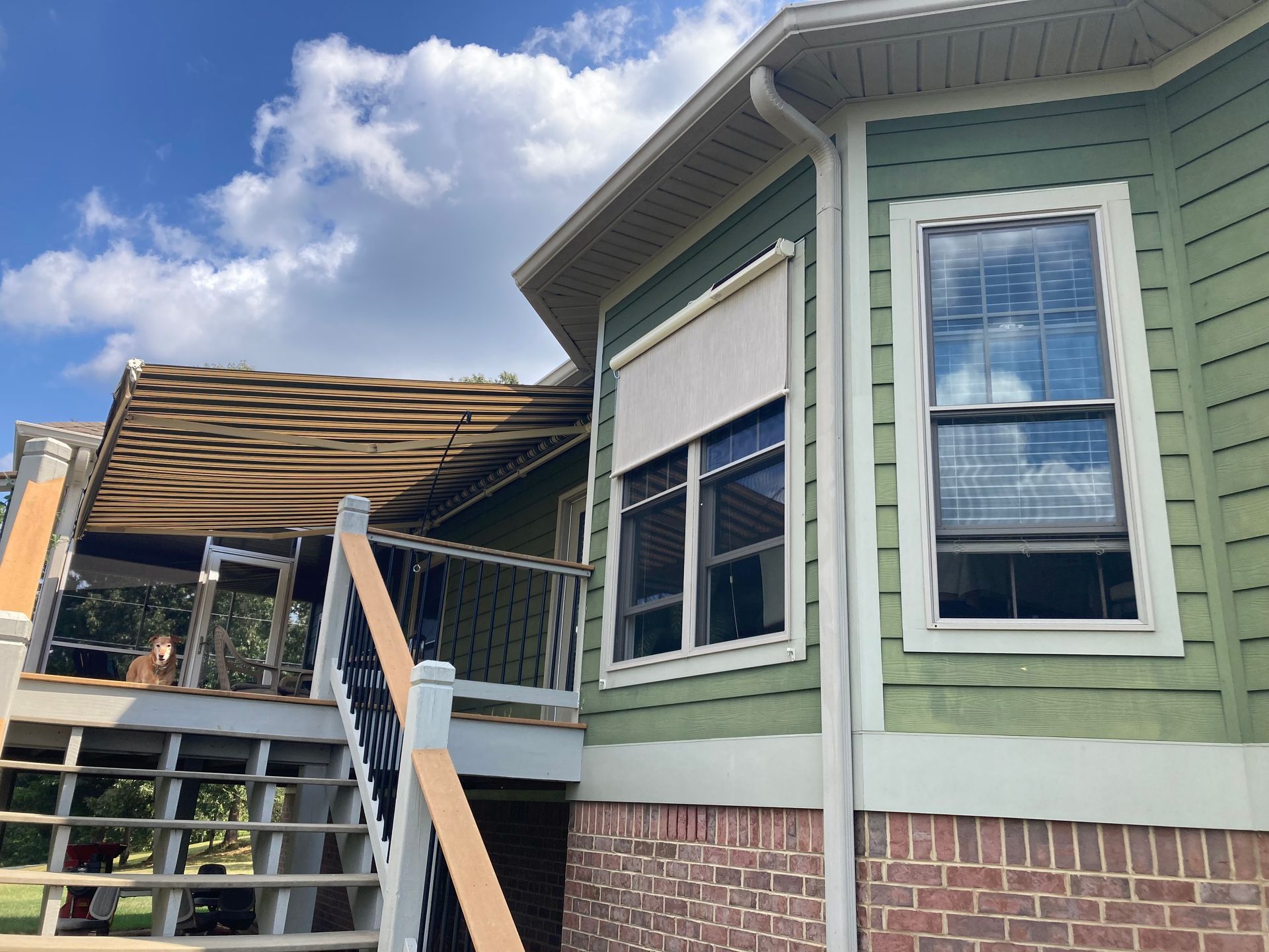 Green house exterior with windows, deck, and a retractable shade. Blue sky with clouds in the background.