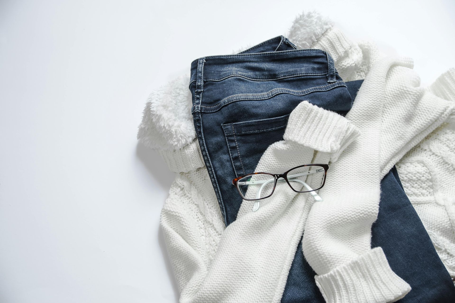 Suéter blanco, jeans azul oscuro y anteojos dispuestos sobre una superficie blanca.