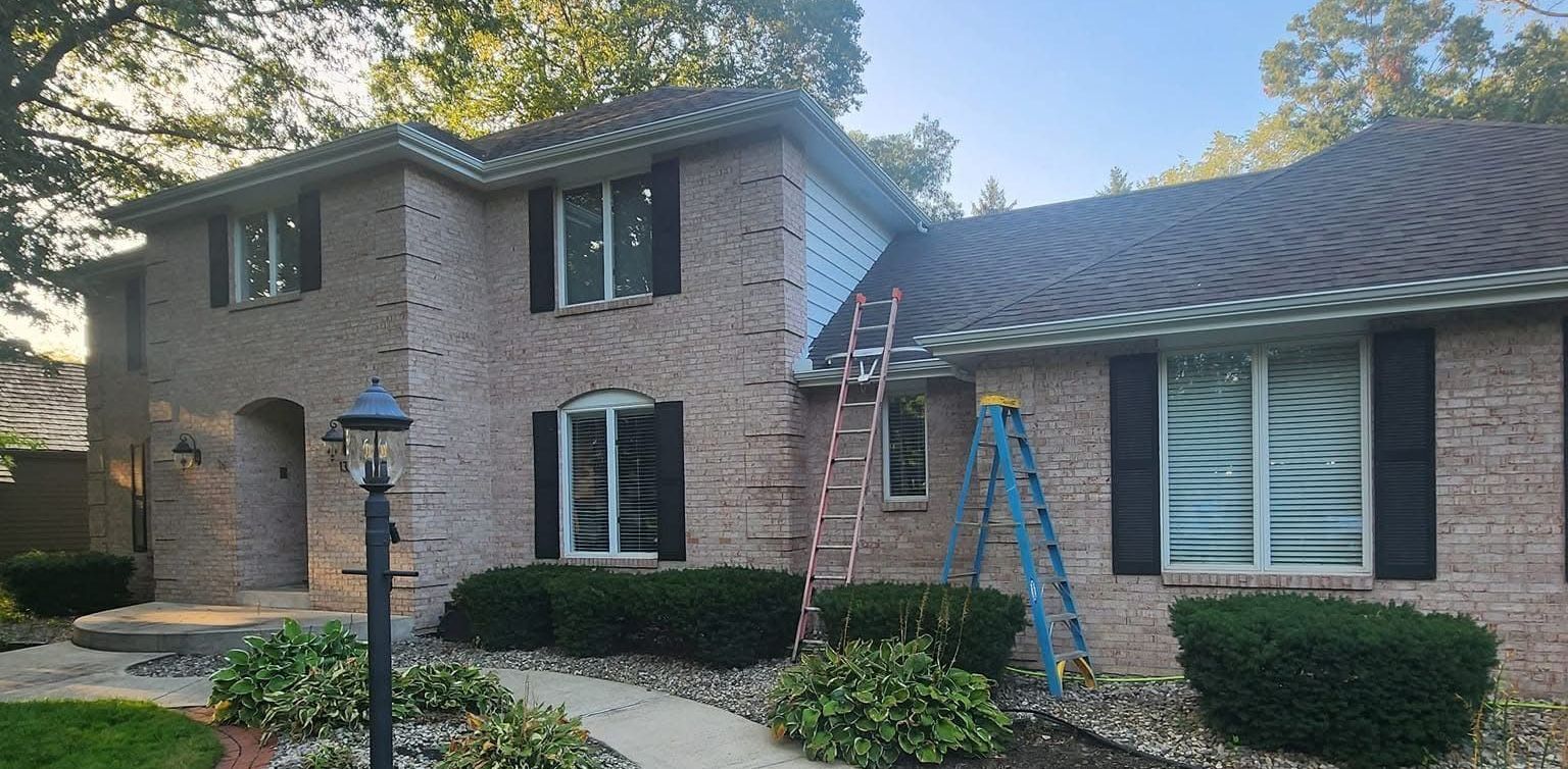 A two-story light-colored brick house with a dark roof and black shutters, featuring a ladder leaning against the siding.