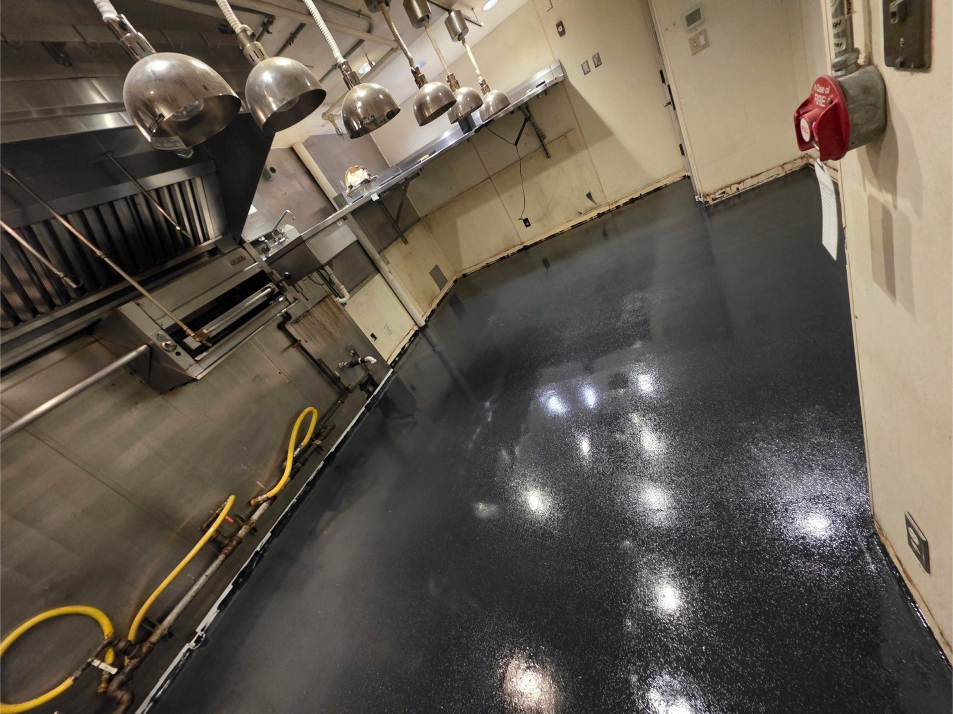 Commercial kitchen floor being pressure washed, with reflective wet concrete and stainless steel equipment.