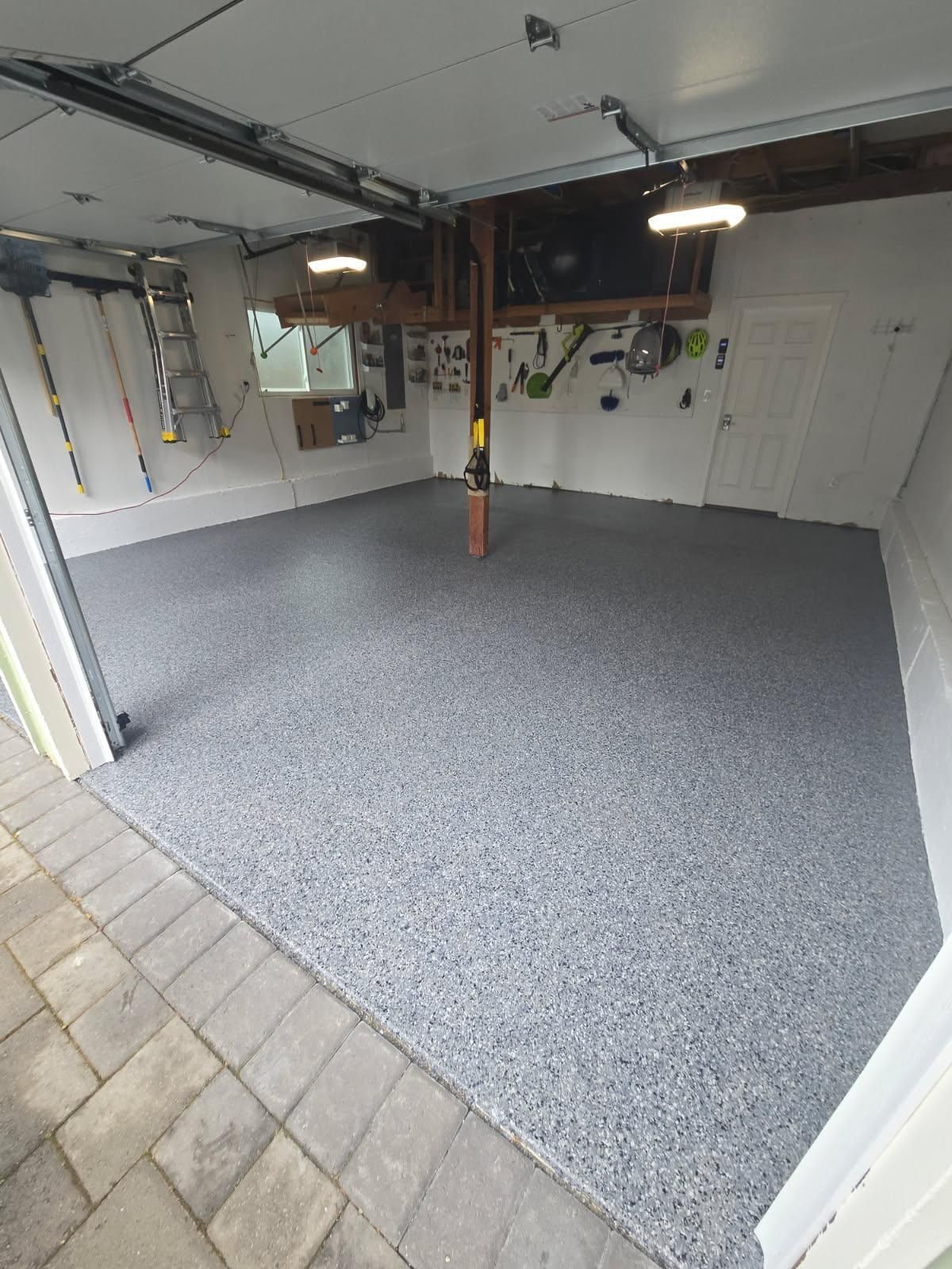Empty garage with gray speckled floor coating, white walls, and overhead lights open to a tiled driveway