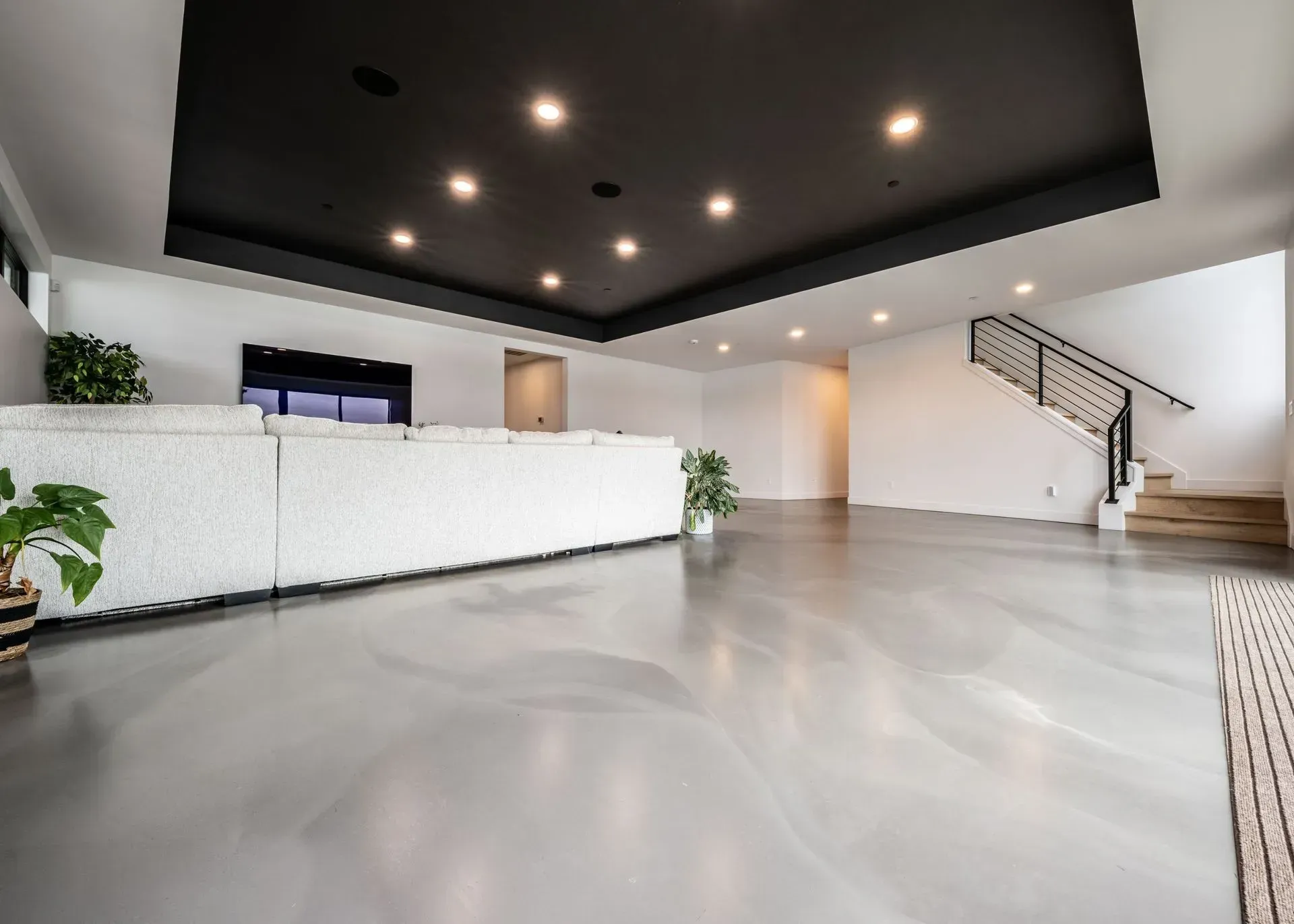 Spacious, modern room with gray epoxy floor, white walls, black ceiling with recessed lights, and staircase.