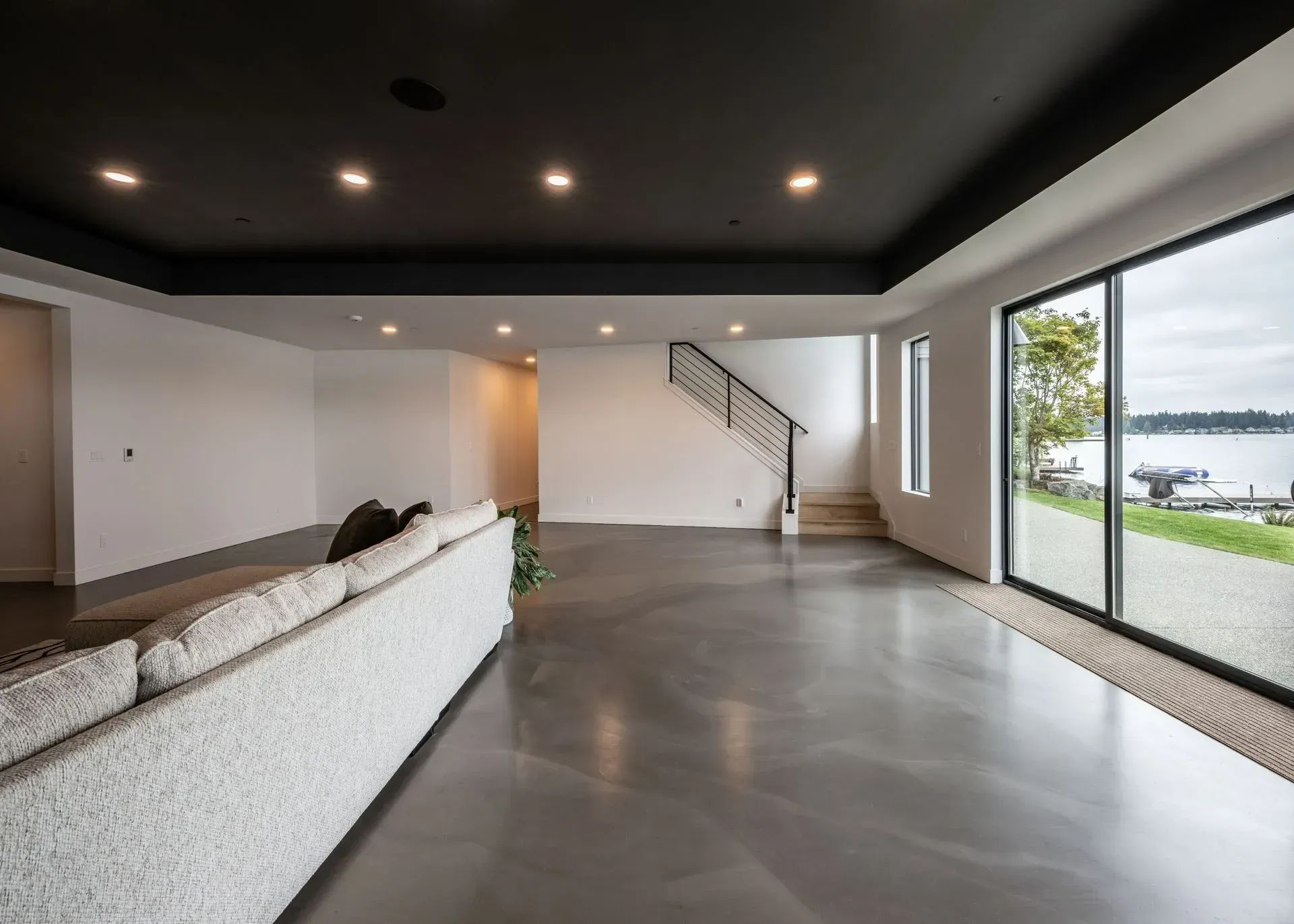 Spacious modern room with dark ceiling, gray flooring, large windows overlooking water, and a white sofa.