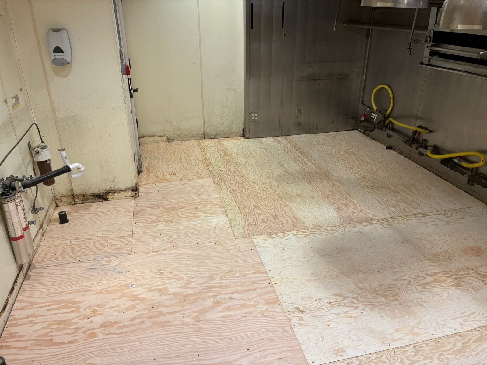 Empty industrial room with plywood-covered floor and beige walls, tools along the right side.