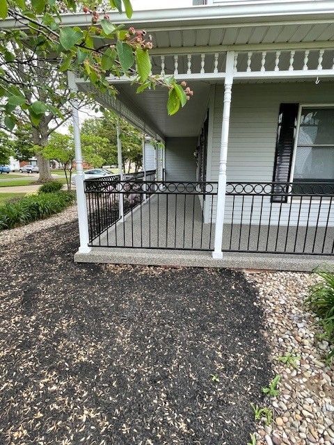Small front porch with black railing, white siding, and a dark mulched garden bed in the yard.