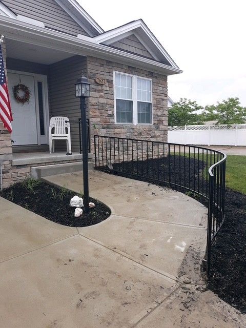 Front porch with stone siding, black railing ramp, and concrete walkway leading to a chair near the entrance