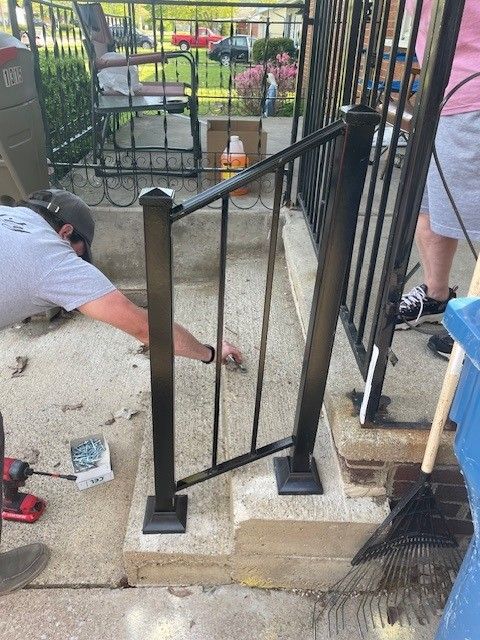 Workers repairing a black metal fence post on a concrete patio beside a house