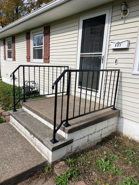 Front porch with black metal railings, beige siding, and a white storm door beside a concrete ramp.