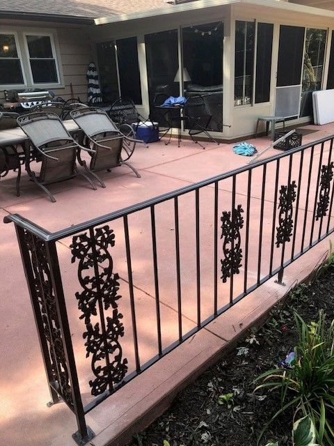 Patio with black metal railing, pink concrete floor, chairs, and house windows and doors beyond