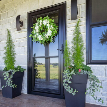 Black front door with a green wreath, flanked by two potted shrubs against a stone facade. Black front door with a green wreath, flanked by two potted shrubs against a stone facade.