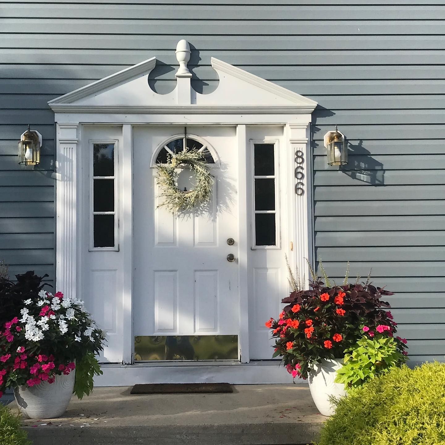A white front door with a wreath, surrounded by potted flowers and porch lights, on a house with light gray siding.