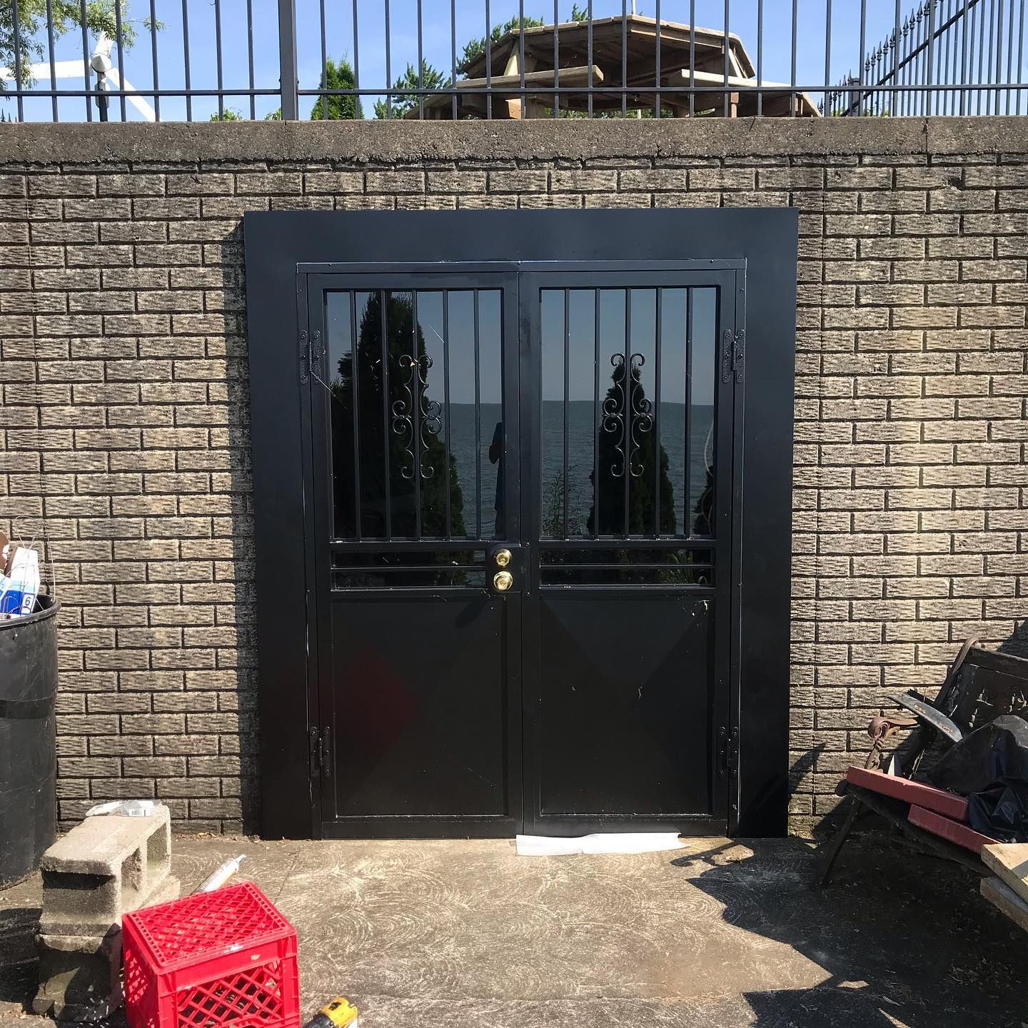 Black double security doors with metal bars and glass panes installed in a textured light brown brick wall.