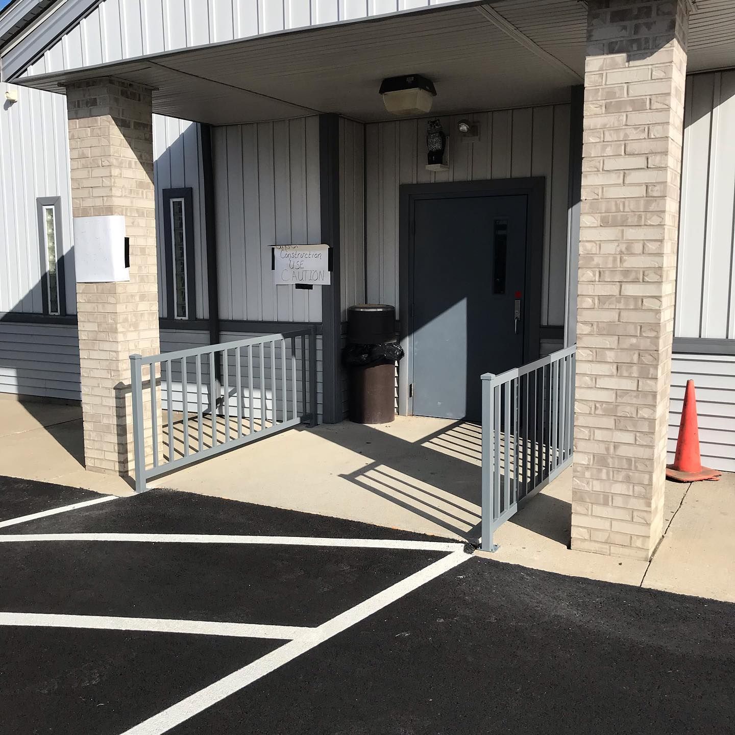 Entrance with a ramp, gray metal door, brick pillars, and a traffic cone on an asphalt parking lot.